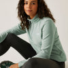 Woman wearing a light green pullover with a brand logo, sitting on the floor against a plain background.
