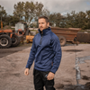 A man wearing a blue quarter-zip pullover and black pants stands in an outdoor farm setting with tractors and a trailer in the background.