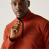 A close-up of a man wearing a rust-orange fleece pullover with a black half-zip and a small black logo on the chest, set against a plain light background.