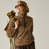 A woman wearing a brown and beige leopard-print fleece pullover and dark sunglasses, holding hiking poles and a backpack against a plain light gray studio background.