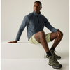 A male model sits on a white rectangular block in a studio setting, wearing a slate blue quarter-zip long-sleeve shirt, light olive green shorts, black socks, and olive green hiking boots.