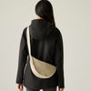 A woman viewed from behind wearing a black hooded jacket and carrying a beige crescent-shaped crossbody bag with a wide strap against a plain light gray studio background.