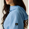 Close-up of a woman wearing a light blue hooded jacket, showing the side profile with a black logo patch on the sleeve.