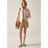 A woman stands in a studio wearing a light pink zip-front hoodie over a white top, tan shorts, and beige slide sandals, while carrying a large tan tote bag.
