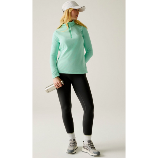 A woman modeling a mint green long-sleeve quarter-zip athletic top, paired with black leggings, a white cap, white socks, and gray running shoes, holding a silver water bottle against a light gray studio background.