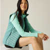 A woman sits on a white block wearing a mint green quilted puffer vest over a matching long-sleeve top, paired with black shorts, against a plain light beige studio background.