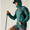 A man wearing a teal and black outdoor jacket, black cap, and hiking backpack with trekking poles, posed against a plain light beige studio background.