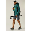 A man wearing a teal hooded hiking jacket, gray shorts, and hiking boots walks while holding trekking poles against a plain light gray studio background.