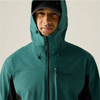 A man wearing a teal-green hooded rain jacket with black side panels and a black cap, photographed against a plain light beige studio background. The jacket has a front zipper, drawcord hood, and a small visible REGATTA brand mark on the left chest.