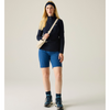 A woman stands against a plain light beige studio background wearing a dark navy long-sleeve quarter-zip top, bright blue fitted shorts, a cream crossbody bag, a dark navy beanie, dark socks, and hiking shoes.