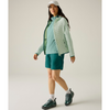 A woman wearing a blue technical jacket, over a dark long-sleeve shirt, light blue shorts, and white athletic socks and running shoes, photographed against a plain light gray studio background.