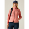 A woman modeling a salmon pink fleece jacket with a black zipper, paired with navy blue pants, a navy blue cap, and a black backpack, set against a plain light beige background.