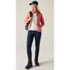 A woman modeling a salmon pink zip-up fleece jacket over a white t-shirt, paired with navy blue leggings, hiking boots, and a navy blue baseball cap, standing against a neutral studio background.