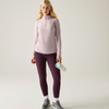 A woman stands against a light gray studio background wearing a light pink long-sleeve quarter-zip athletic top and dark purple leggings, with white socks and gray sneakers, and holding a white water bottle.